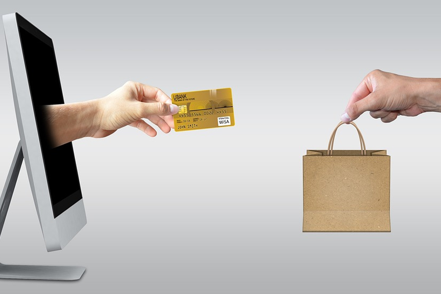 two computers facing each other, with a hand holding a credit card reaching out of one screen and a hand holding a shopping bag reaching from the other